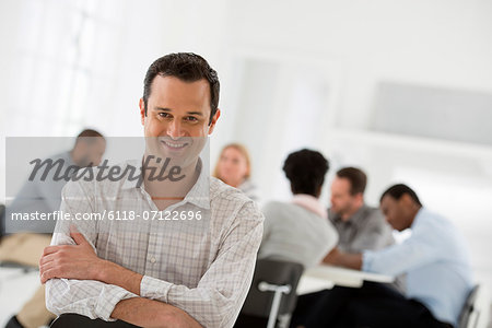 Office Interior. A Man Seated Separately From A Group Of People Seated Around A Table. A Business Meeting.