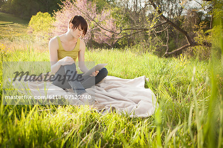 A Young Woman Sitting In A Field, On A Blanket, Reading From A Digital Tablet.