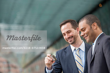 City. Two Men In Business Suits, Looking At A Smart Phone, Smiling.