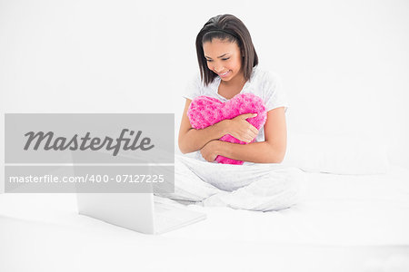 Focused young dark haired model holding a pillow and looking at a laptop in bright bedroom