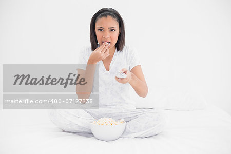 Focused young dark haired model watching tv and eating popcorn in bright bedroom