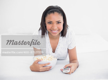 Pleased young dark haired model eating popcorn while watching tv in bright bedroom