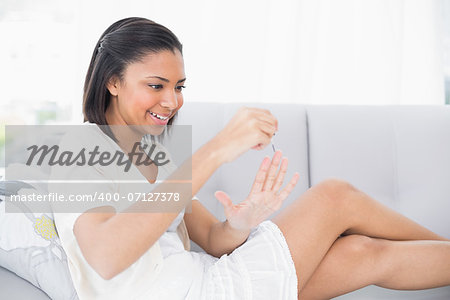 Charming young dark haired woman in white clothes applying nail polish in a living room