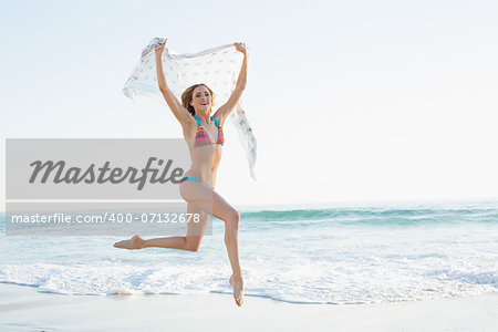 Gleeful slender woman jumping in the air holding shawl on the beach