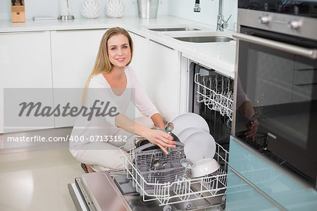 Smiling gorgeous model kneeling behind dish washer in bright kitchen