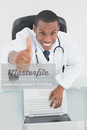 Overhead portrait of a smiling male doctor with laptop gesturing thumbs up at medical office
