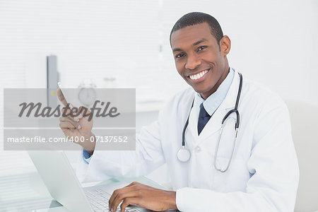 Portrait of a smiling male doctor text messaging while using laptop at medical office