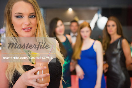 Blonde woman standing in front of her friends holding cocktail and looking at camera
