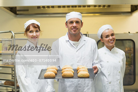 Young male baker holding a baking tray with rolls on it posing with colleagues