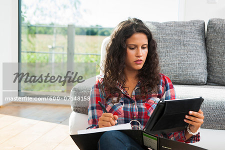 Teenage girl sitting on floor next to sofa, writing in binder and using tablet computer, Germany