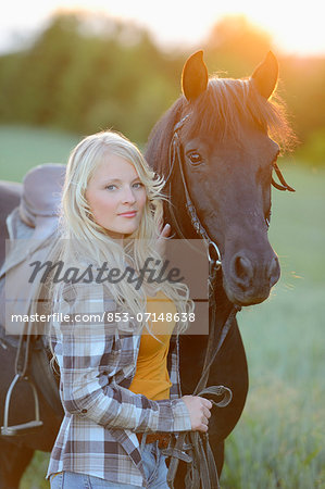 Young woman standing beside a horse on a meadow at sunset