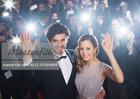 Celebrity couple waving on red carpet with paparazzi in background