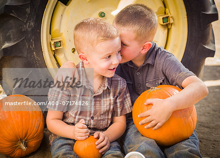 Two Boys Sitting Against a Tractor Tire Holding Pumpkins and Whispering Secrets in Rustic Setting.