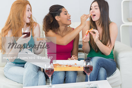 Happy young female friends eating pizza with wine on sofa at home