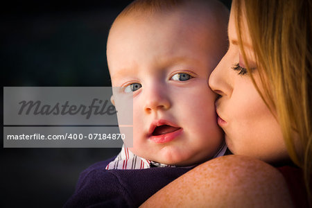 Adorable Red Head Infant Boy is Kissed By His Mother Outdoors in Dramatic Lighting.
