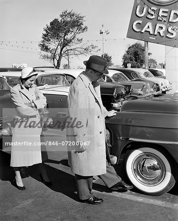 1950s COUPLE IN USED CAR LOT MAN KICKING TIRE
