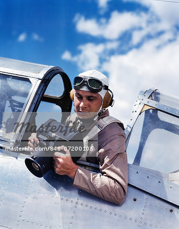 1940s ARMY AIR CORPS MAN SITTING AIRPLANE COCKPIT WEARING FLYING GOGGLES HELMET PARACHUTE TAKING PHOTOGRAPHS WITH AERIAL CAMERA