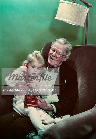 1940s 1950s GRANDDAUGHTER SITTING ON SMILING GRANDFATHER'S LAP TALKING ON TOY TELEPHONE