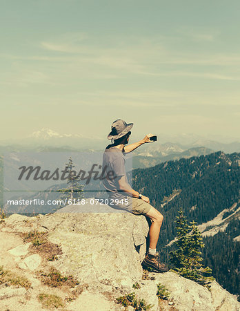 A hiker on a mountain summit, holding a smart phone, at the top of Surprise Mountain, in the Alpine Lakes Wilderness, in Mount Baker-Snoqualmie National Forest.