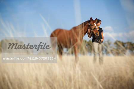 A woman leading a horse on the halter through the long grass in a field.