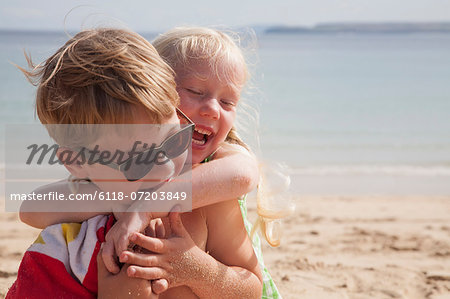 A brother and sister play fighting on the beach. A boy in sunglasses and a younger girl with her arms around his neck.