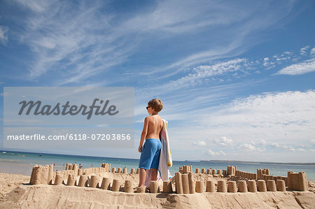 A boy standing beside a sandcastle, on top of a mound of sand. Beach.