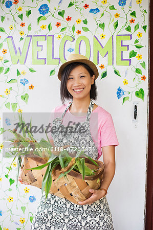 Working on an organic farm. A young Asian woman by the Welcome sign with a large basket of vegetables, freshly picked.