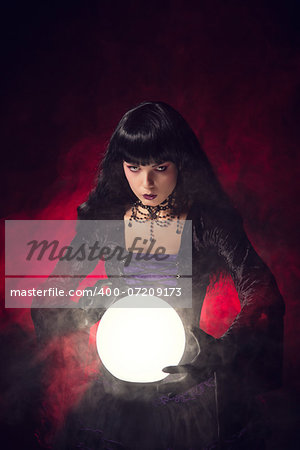 Beautiful gothic style fortune teller with a crystal ball, studio shot over smoky background