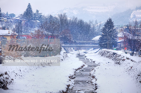 Banks of small creek, bridge and private houses among trees covered by snow in Alba, northern Italy.