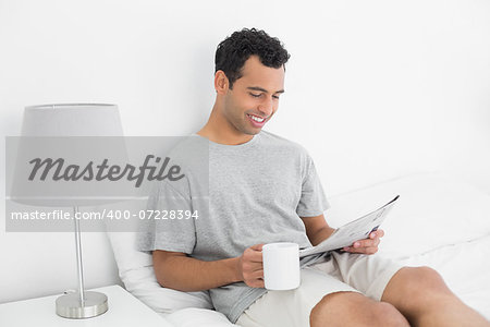 Relaxed young man reading newspaper while holding coffee cup in bed at house