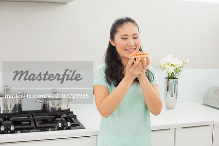 Smiling young woman eating a slice of pizza in the kitchen at home