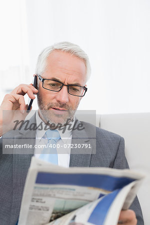 Elegant mature businessman using cellphone while reading newspaper on sofa in living room at home