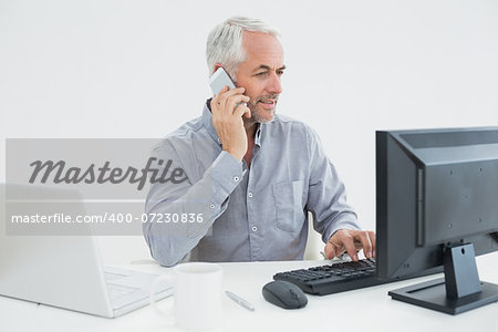 Mature businessman with cellphone, laptop and computer at desk against white background