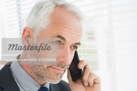 Serious mature businessman peeking through blinds while on call in office