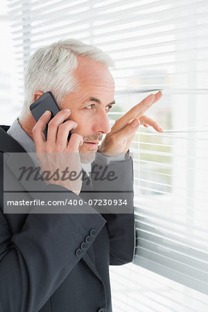Serious mature businessman peeking through blinds while on call in office