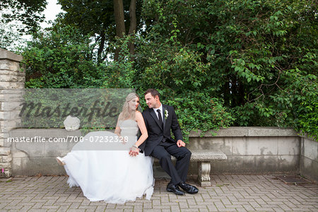 Bride and Groom sitting on stone bench outdoors, holding hands and looking at each other on Wedding Day, Canada