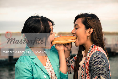 Young women biting opposite ends of sandwich