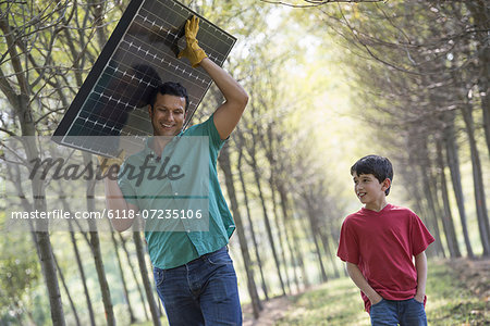 A man carrying a solar panel down an avenue of trees, accompanied by a child.