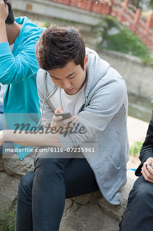 Young man in a gray hooded sweatshirt looking down at his phone and texting outdoors