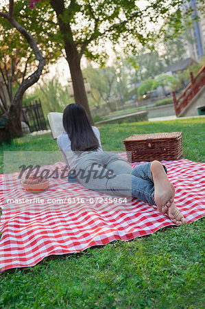 Young woman lying on her stomach on a checkered blanket and reading in the park, having a picnic