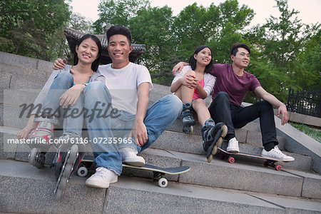 Two young couples sitting and resting on concrete steps outside with skateboards and roller blades