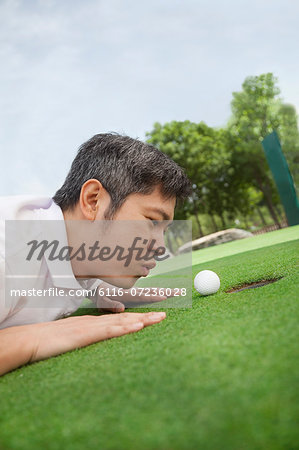 Mid adult man lying down in a golf course trying to blow the ball into the hole