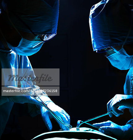 Two surgeons looking down, working, and holding surgical equipment with patient lying on the operating table