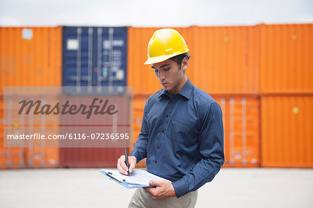 Smiling young engineer in protective work wear in a shipping yard examining cargo and writing on clipboard