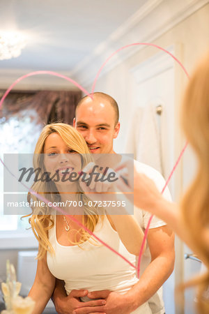 Young couple embracing and looking in bathroom mirror together, drawing a heart with lipstick on mirror, Canada