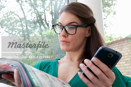 Young woman wearing glasses reading magazine holding cell phone