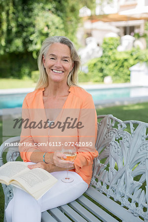 Smiling senior woman reading book and drinking wine in backyard