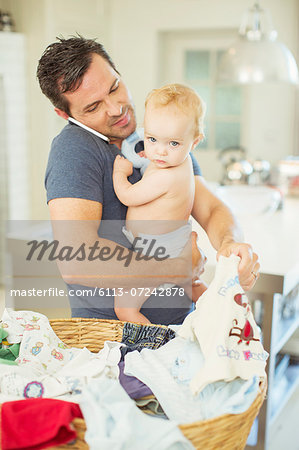 Father holding baby and folding laundry while talking on cell phone