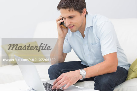 Smiling young man using laptop and cellphone in the living room at home