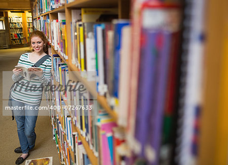 Cheerful student reading book leaning on shelf in library at the university